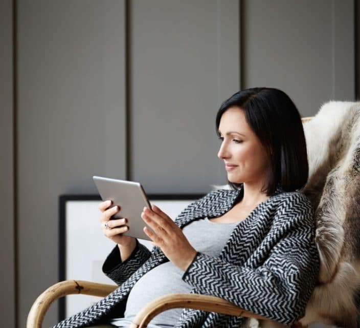 Pregnant woman using a digital tablet while relaxing on a rocking chair at home